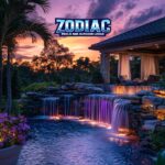 A stunning tropical backyard pool by Zodiac Pools & Outdoor Living, featuring cascading rock waterfalls illuminated by colorful LED lights at sunset, surrounded by palm trees, lush landscaping, and a cozy covered lounge area.