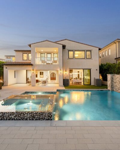 A stylish two-story home with a contemporary backyard featuring a rectangular swimming pool and an elevated spa with a stone spillway, surrounded by a clean patio design and warm evening lighting.